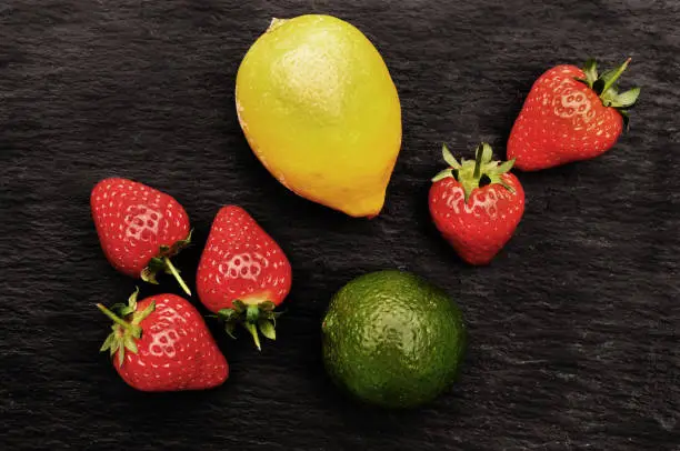 A arrangement of fruit including a yellow lemon, a green lime and five red strawberries on dark black grey textured slate. Flat lay from above