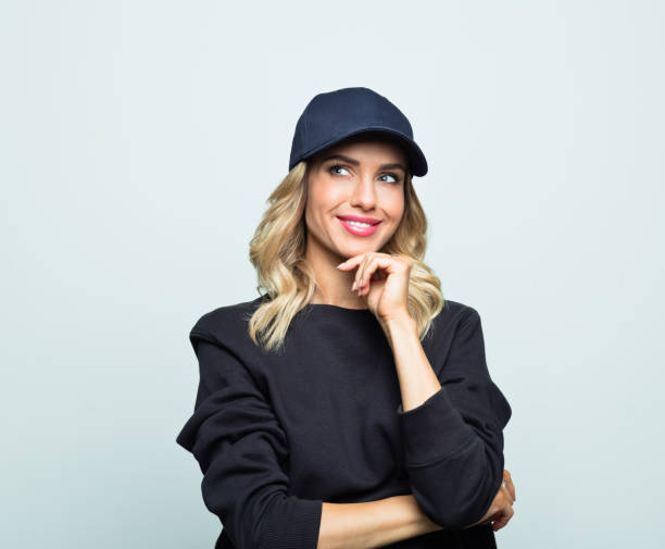 Portrait of cheerful young woman Portrait of smiling young woman looking away with hand on chin. Modern beautiful female is wearing street style black blouse and baseball cap. She is standing against grey background. woman wearing baseball cap stock pictures, royalty-free photos & images
