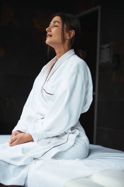 istock Beautiful elegant Caucasian young lady in white plush bathrobe sitting on massage table in room indoors 1316811384