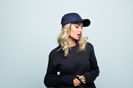 Modern beautiful female is wearing street style black blouse and baseball cap. She is standing against grey background.