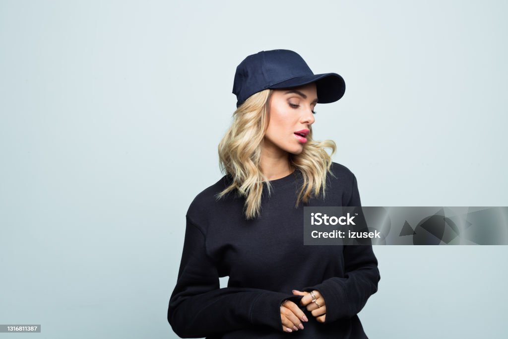 Portrait of pensive young woman Modern beautiful female is wearing street style black blouse and baseball cap. She is standing against grey background. Women Stock Photo