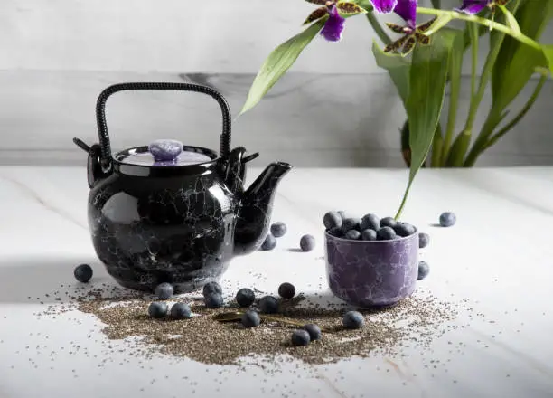 still life with a black ceramic teapot and a cup of tea with blueberries on a light background with an orchid, chia seeds and berries are scattered on the table