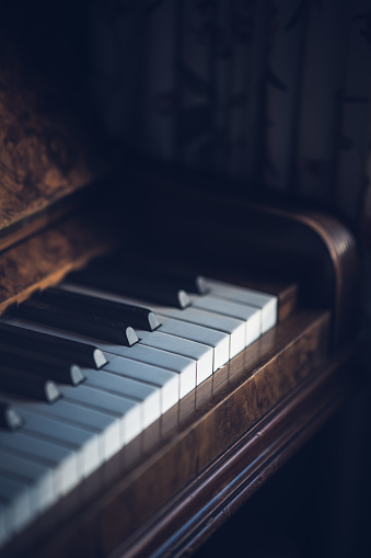 Old vintage mahogany acoustic upright piano with subtle sunlight shining on piano keys, with selective focus and dark retro colour grading