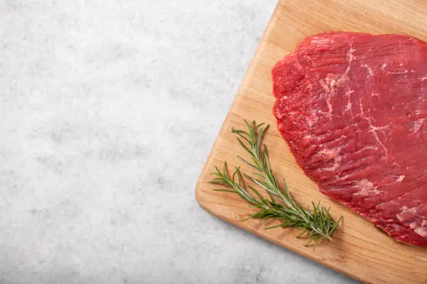 A raw beef flank steak on butcher cutting board sitting on white marble with copy space.