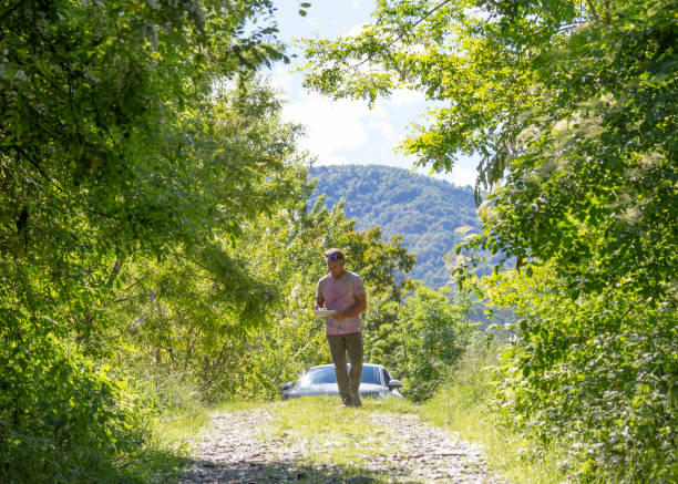 Man gets out of car and relaxes on sunny mountain road He looks at tablet for directions and walks through lush forest environment computer cloud leadership stock pictures, royalty-free photos & images