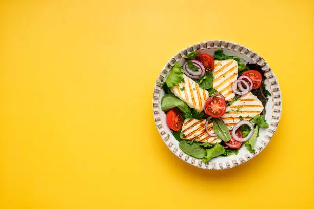 Healthy green salad with grilled halloumi cheese on yellow background, top view, space for text