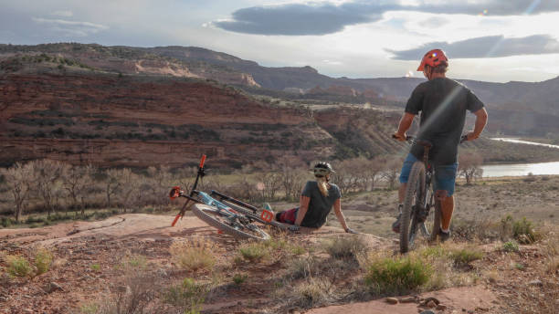 Mountain biking couple relax on mountain ridge Man makes his way over to woman sitting down, Colorado River and desert landscape below fruita colorado stock pictures, royalty-free photos & images
