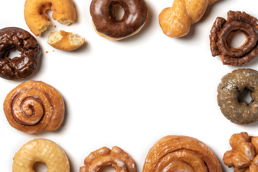 Overhead horizontal photograph of an assortment of donuts on a white background. Glazed, cake, and chocolate covered donuts as well as a cinnamon roll and specialty donuts are arranged in a border pattern.
