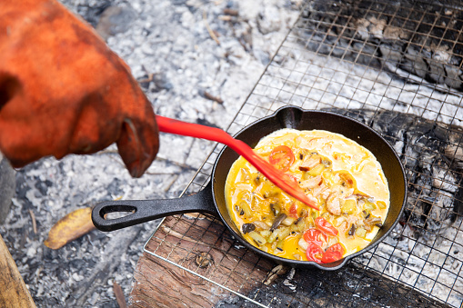 A close up of a hand in glove with a spatula cooking an omelet over a camp fire.