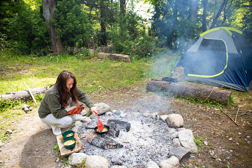 A Japanese lady cooking an omelette over hot coals of a camp fire in an iron skillet at her camp site in the wilderness by her self.