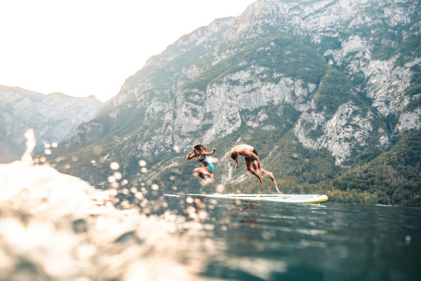 Vacationing Couple Jumping Off Paddleboard into Lake Bohinj Surface level view of energetic mid adult couple jumping off paddleboard into lake while on summer vacation in Triglav National Park. paddleboard surfing water sport low angle view stock pictures, royalty-free photos & images