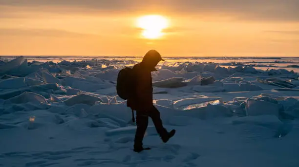 Photo of Silhouette of tourist walking on ice surface with ice hummock rising above the frozen lake of Baikal at sunrise.