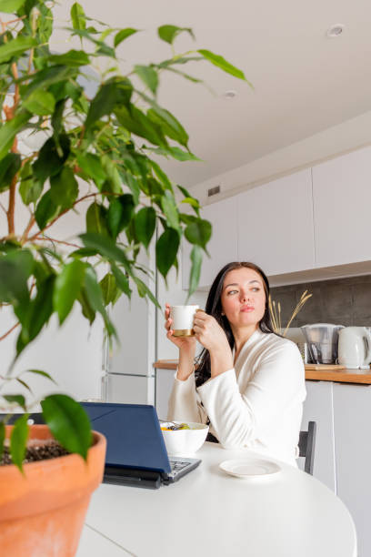 istock Young woman in comfortable white robe works on laptop while sitting at table the kitchen. Work at home for breakfast. 1316897110