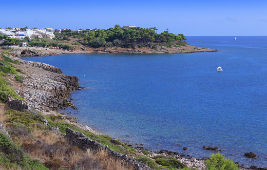 Wide-angle view of the idyllic northernmost cove of Cala Tarida, on the western coast of Ibiza. A perfectly clear sky, the dazzling bright light of a Mediterranean summer noon, colourful cliffs dotted with Mediterranean scrub, a few sailing boats and motorboats rocking gently on transparent waters, sparse bathers engaged in different activities and enjoying a privileged environment. The small group of fishermen’s huts of Es Pujolets can be seen on the background, the peculiar rock of Escull des Camió at the centre of the image. High level of detail, natural rendition, realistic feel. Developed from RAW.