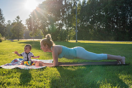Mother exercising while her little baby boy sitting on blanket in the park