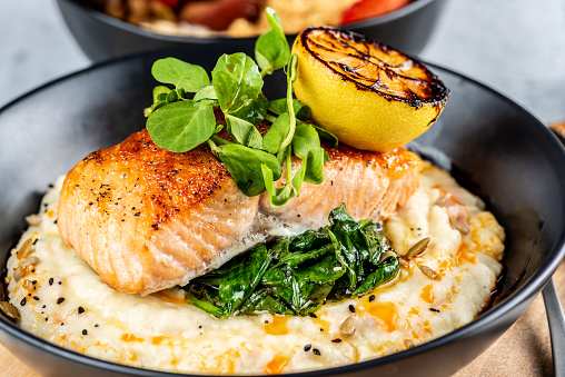 A seafood dinner serve of a piece of Salmon on top of mashed potatoes, in a dark round plate