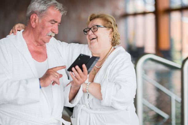 glückliches senior-paar mit handy auf dem badeanzug in bademänteln und umarmung - overweight smiling senior women 70s stock-fotos und bilder