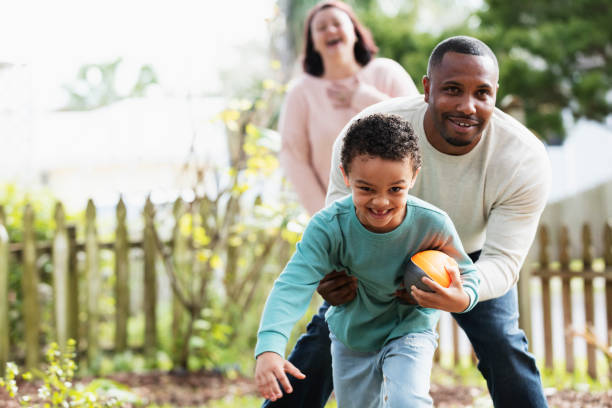 Boy, father playing with football in yard, mom laughing A little boy having fun playing with his father in the backyard. He is holding a football, trying to run away from dad. The African-American man is in his 30s. His son, 5 years old, is mixed race African-American and Caucasian. They are smiling at the camera. Mom is out of focus in the background, laughing. common couple men outdoors stock pictures, royalty-free photos & images