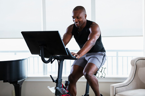 A mid adult African-American man in his 30s exercising at home in his living room, on an exercise bike. He is an athlete with a muscular build.