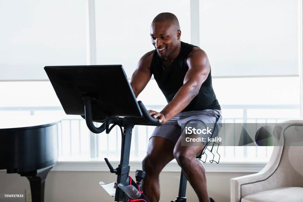 African-American man on exercise bike at home A mid adult African-American man in his 30s exercising at home in his living room, on an exercise bike. He is an athlete with a muscular build. Exercise Bike Stock Photo