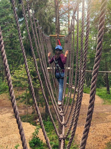 A girl crossing a rope bridge of an obstacle course in a forest wearing safety equipment.
