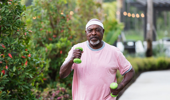 A senior African-American man in his 70s exercising outdoors. He is power walking or jogging, with dumbbells in his hands. He is looking at the camera with a serious expression.