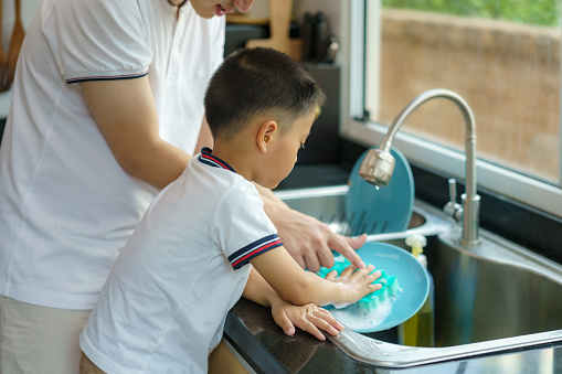 Asian father is teaching his son how to wash dishes, help with housework in the kitchen at home, fathers interact with their children throughout the day.