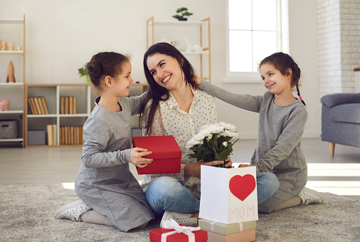 Two little daughters greet their mother and give her flowers, a gift and a postcard. Mom and children sitting on the floor at home smiling and hugging. Concept of family celebration and togetherness.