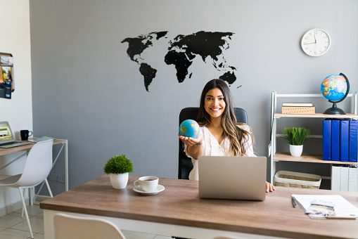 Holding the world in my hands. Attractive female travel agent showing a globe and smiling while working at her modern office
