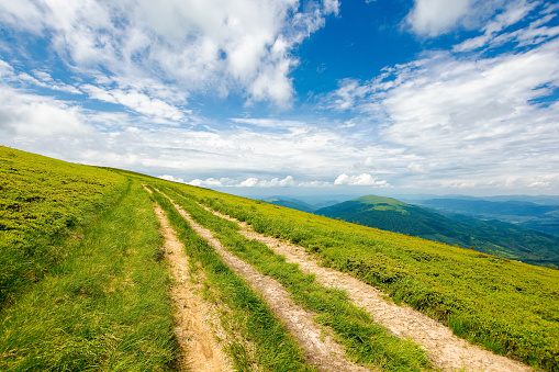 country dirt road through grassy hillside. mountain ridge in the distance beneath a gorgeous cloudscape on the blue sky. travel backcountry concept