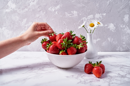 Woman takes ripe strawberry from white ceramic bowl on gray concrete background, copy space. Healthy food concept, still life