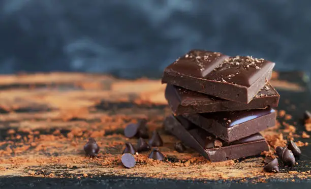 Photo of Chocolate pieces stack with chocolate flakes, drops and cocoa powder on dark background