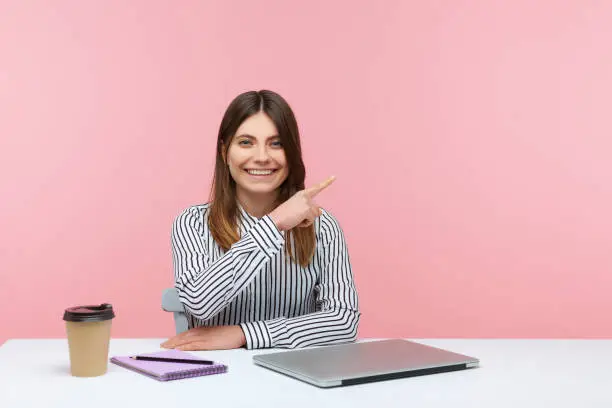 Smiling positive woman advertising agency worker pointing finger away paying attention and showing freespace for your advertising sitting at workplace. Indoor studio shot isolated on pink background