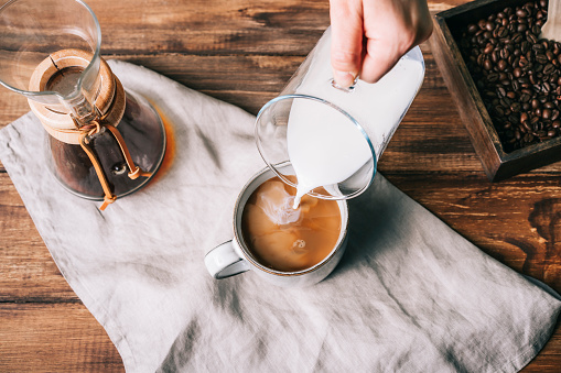 Pouring milk with a glass carafe into a cup with coffee on a brown wooden table