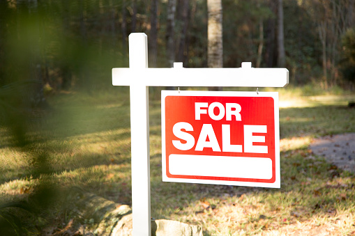 Home for sale with red and white real estate sign during the fall season.  Fall season with leaves on ground.  Front porch and windows in background.  Residential neighborhood.  Moving house, relocation concept.  Lot for sale.  Land grab in Texas.