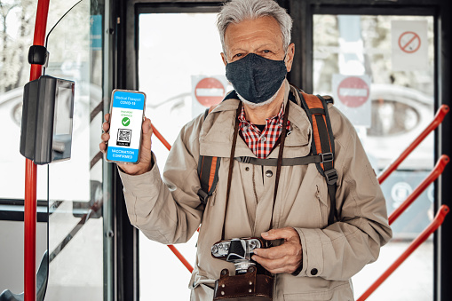 Handsome senior tourist man with black protective face mask using city transport vehicle. He is holding smart phone and showing electronic Covid-19 immunity passport. Coronavirus and tourism concept.
