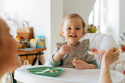 Cheerful baby girl sitting on high chair while eating meal. Female toddler is being fed by mother. They are at home.