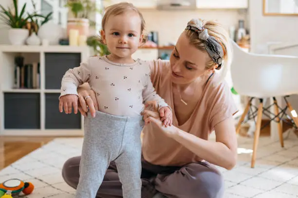 Young beautiful woman assisting daughter in wearing leggings. Portrait of cute baby girl getting dressed by mother. They are in living room at home.