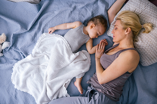 Directly above shot of young woman sleeping with baby daughter. Cute toddler girl is lying by mother on bed. They are relaxing comfortably at home.