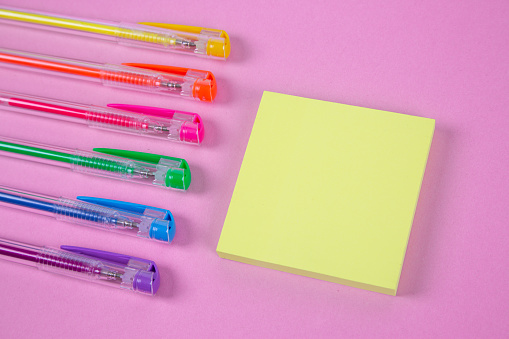 Diagonal view of vibrant colored pens and yellow adhesive note paper on the pink background.
