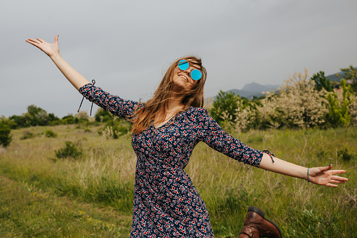 One beautiful young woman in floral dress and sunglasses with raised arms enjoying springtime vacation in nature