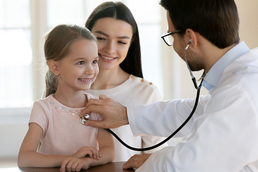 Young male pediatrician hold stethoscope listen to happy small kid girl heart at checkup visit at hospital with mom. Caring man doctor use phonendoscope check lungs breath of little kid in clinic.
