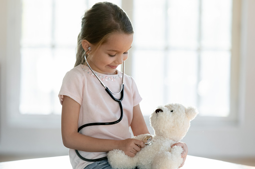 Small Caucasian girl child hold stethoscope listen to fluffy toy heart breath act as doctor. Cute little kid use phonendoscope cure check examine teddy bear, play hospital. Career, healthcare concept.