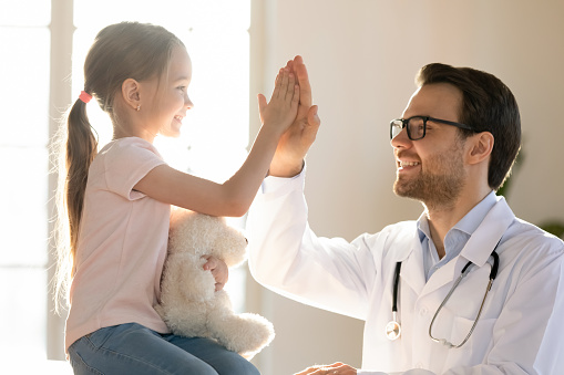 Happy young Caucasian male pediatrician give high five to small kid patient in private hospital. Smiling man doctor or GP make deal with little girl child in clinic. Pediatric, healthcare concept.