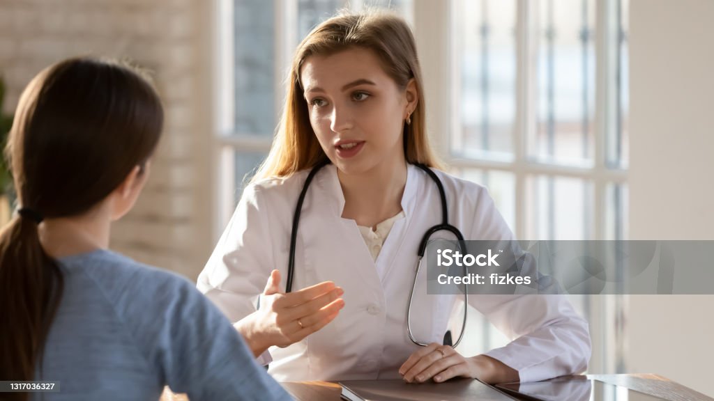 Focused young beautiful female doctor talking with patient. Focused young beautiful female doctor talking with woman at checkup meeting, explaining medical insurance benefits or fiving advice on health treatment procedures, sitting at table in clinic office. Patient Stock Photo