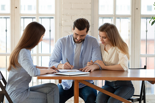 Happy young clients signing paper contract with professional broker or Real Estate Agent, after discussing agreement details. Confident saleswoman watching millennial couple putting signature, purchasing house.