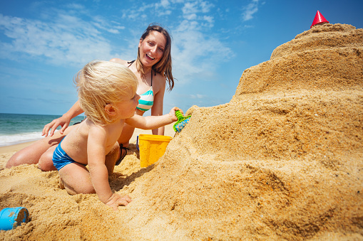 Blond toddler boy play building sand castle on the beach and happy mother laugh on background