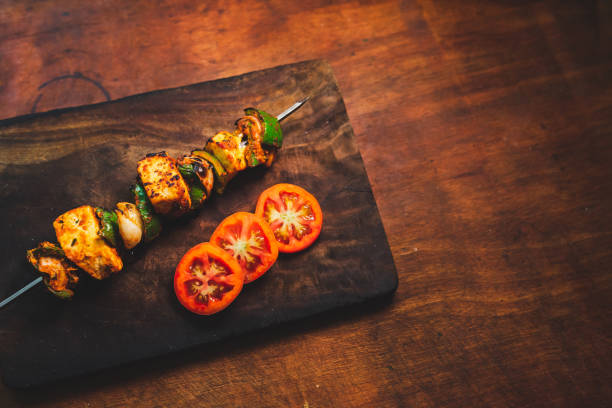 Paneer tikka masala appetiser food served on a barbecue skewer. Cooked Paneer tikka on a skewer with fresh tomatoes and vegetables. Spicy Indian cottage cheese on a dark wooden plate at restaurant table with copy space for text. appetiser stock pictures, royalty-free photos & images