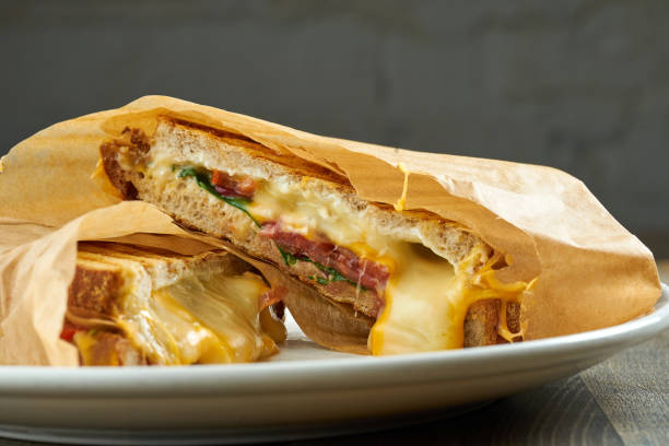 istock Large fried sandwich with ham, tomatoes, pickles and melted cheese, served in parchment on a white plate on a wooden background 1317064839