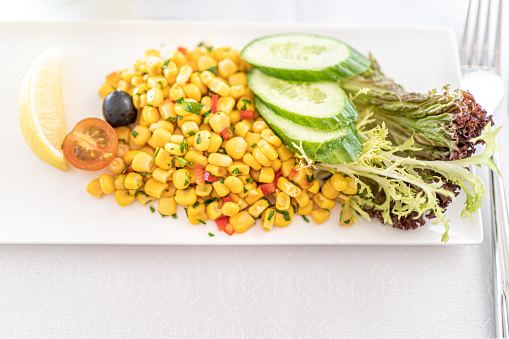 Fresh corn salad on a white plate serves with cucumber, black olive, tomatos, and a slice of lemon